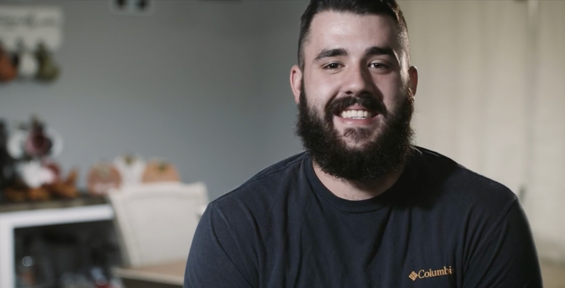 A man with a beard and dark hair smiles while wearing a black Columbia t-shirt. He sits indoors with a blurred background that includes light-colored furniture and decorations.