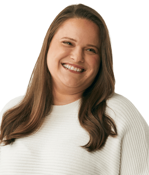 A woman with long brown hair, wearing a white ribbed sweater, smiles warmly while looking slightly to her left. The background is plain and light-colored.