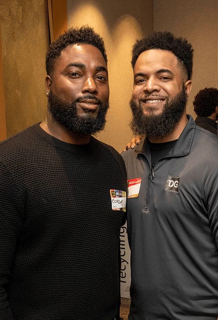 Two men with beards pose together indoors, smiling. Both wear name tags and casual clothing—a black sweater and a gray pullover. The background is softly lit and neutral.