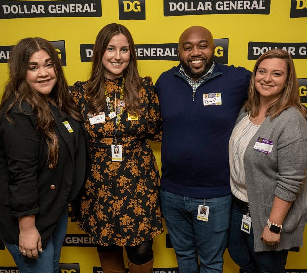 Four people stand smiling in front of a yellow Dollar General backdrop, each wearing ID badges and casual-professional attire.