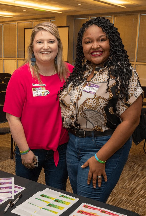 Two women smiling, standing side by side in a conference room with tables, papers, and name tags visible. Both are wearing casual clothes and appear happy, with bright lighting and empty chairs in the background.