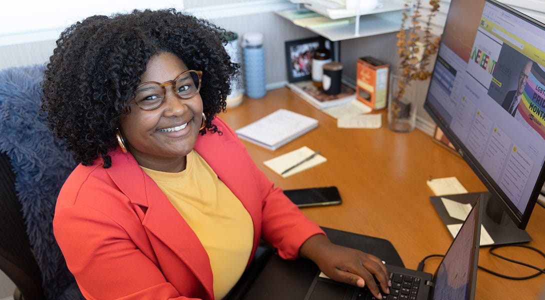 A woman in a red blazer and yellow shirt smiles while working on a laptop at her office desk, with papers, a phone, and a large computer monitor nearby.