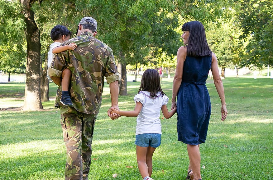 A man in camouflage holds a child and walks hand-in-hand with a girl and a woman through a sunny park, their backs to the camera, surrounded by green grass and trees.