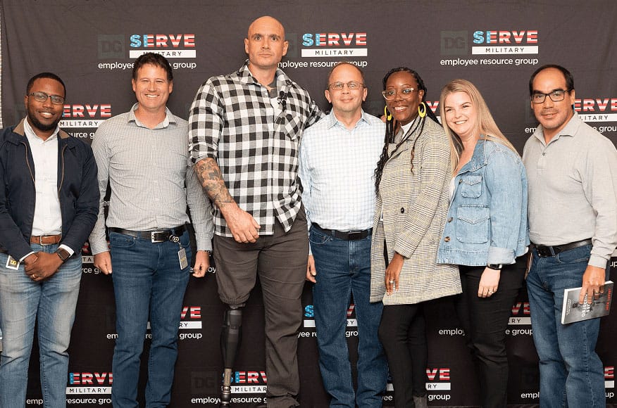Seven people stand together smiling in front of a “DC Serve Military Employee Resource Group” backdrop. One man in the center has a prosthetic leg and is notably taller than the others.