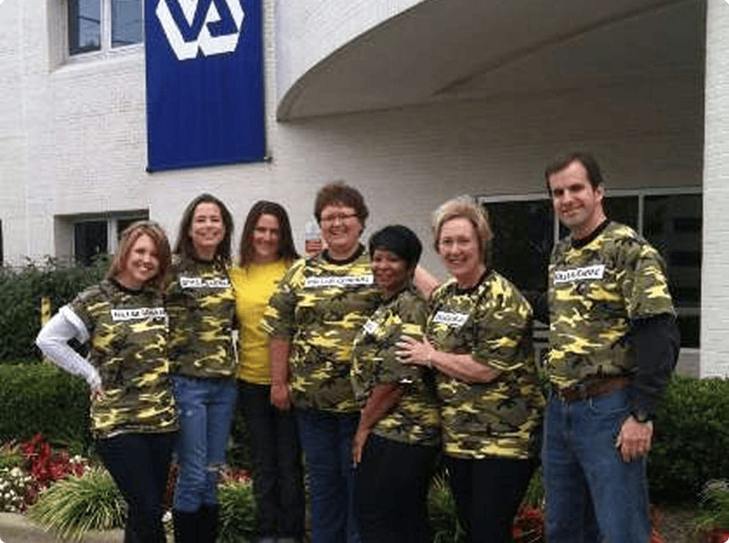 Seven people stand outside a building with a VA sign. Six wear yellow camo shirts and one wears a yellow shirt. They are smiling and posing together near some bushes and flowers.