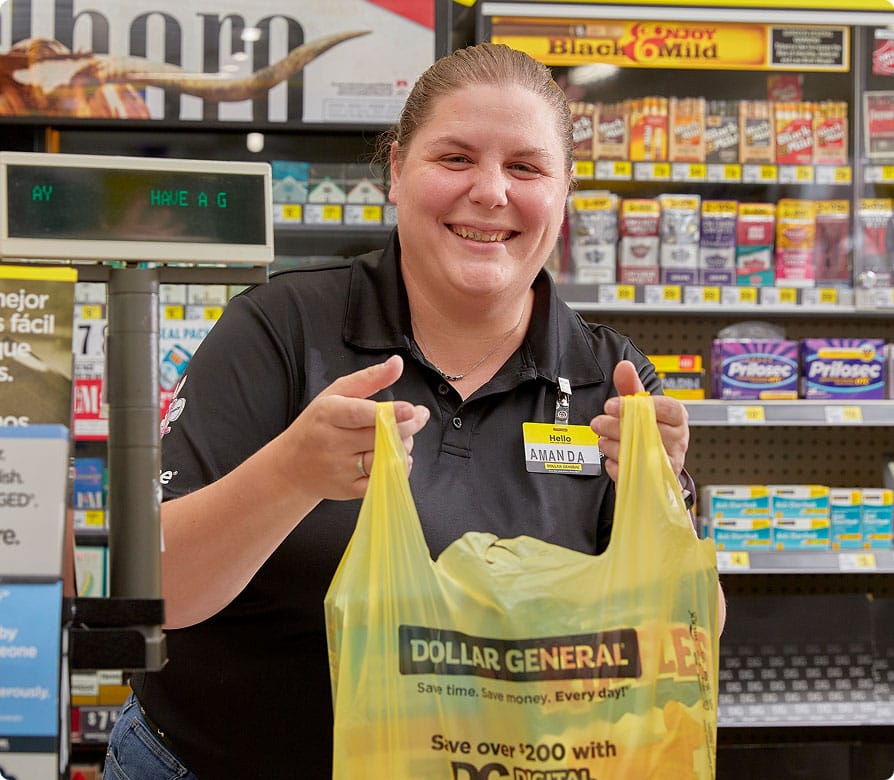 A smiling Dollar General employee holds a yellow shopping bag toward the camera while standing at the checkout counter, with shelves of cigarettes and other products in the background.