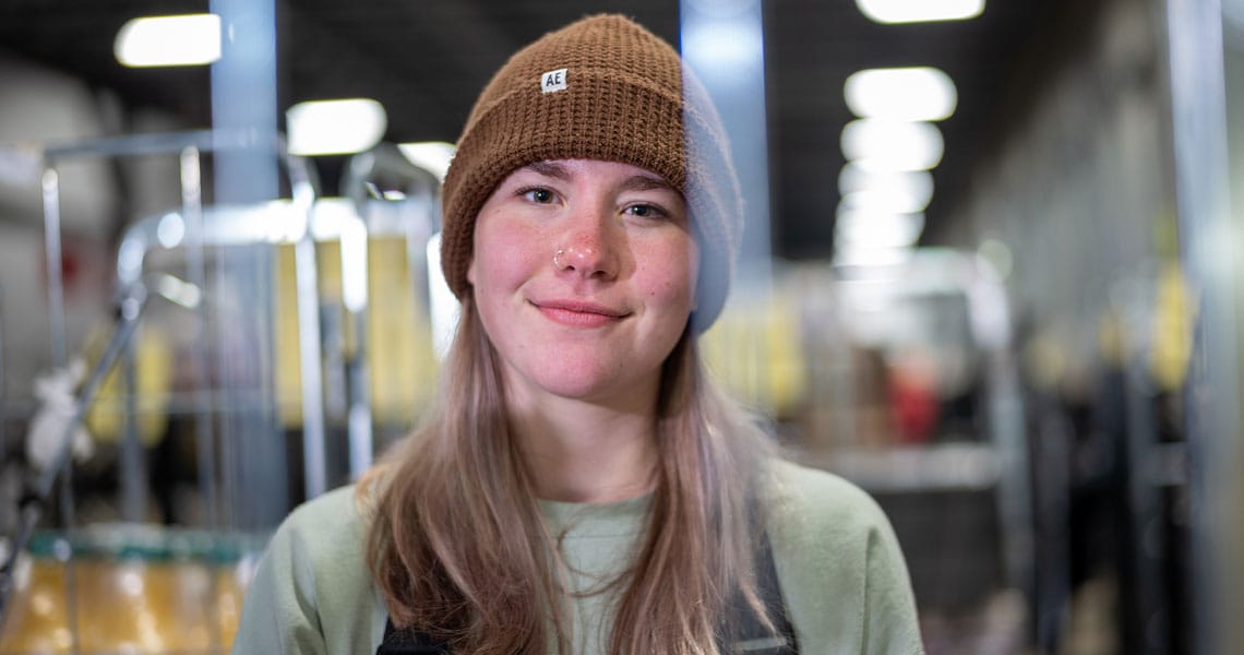 A person with long, light hair wearing a brown knit beanie, green shirt, and black overalls smiles in a brightly lit indoor setting, with blurred shelves and lights in the background.
