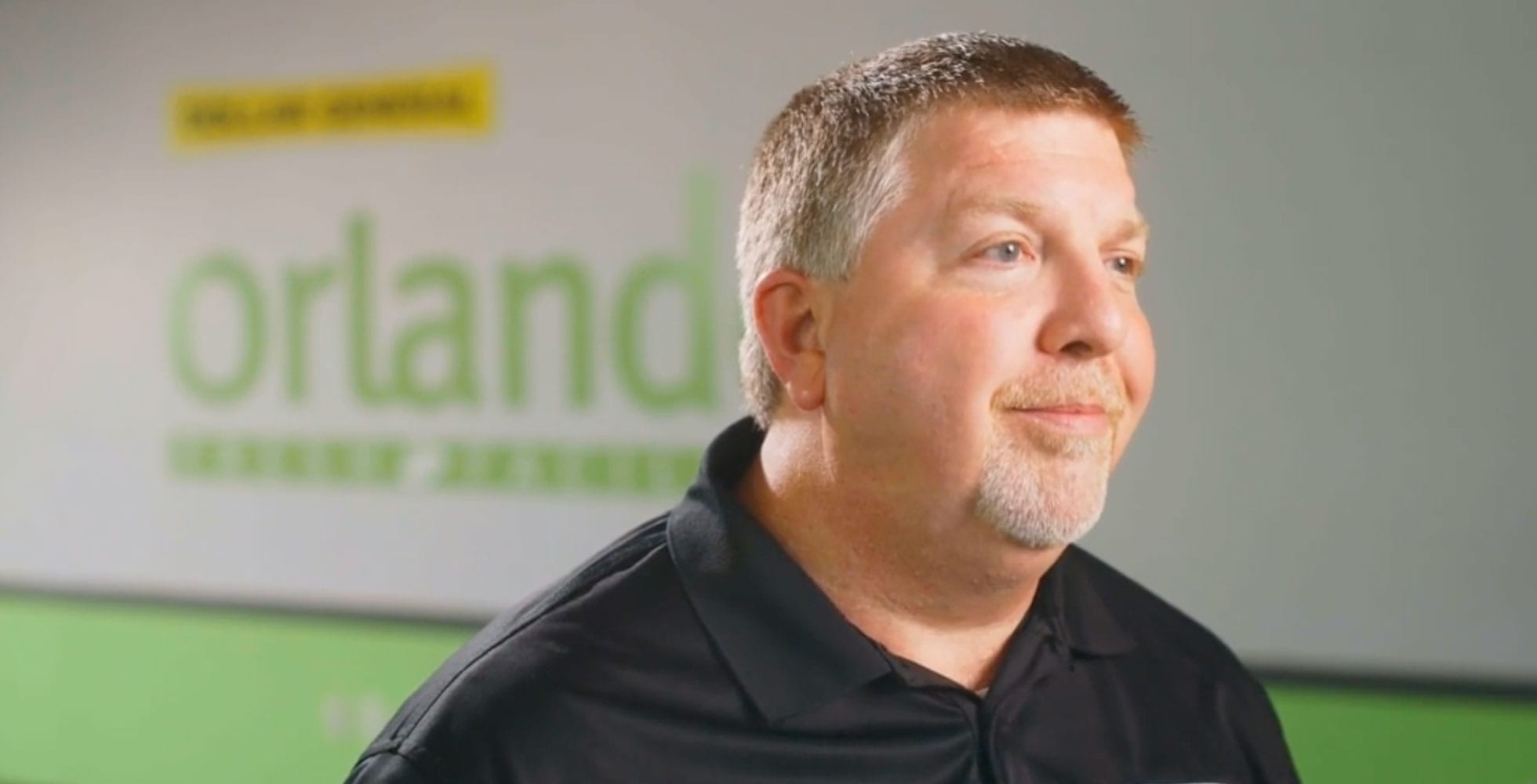 A man with short hair and a goatee, wearing a black polo shirt, stands in front of a blurred wall with green text that partially reads “orland.”.