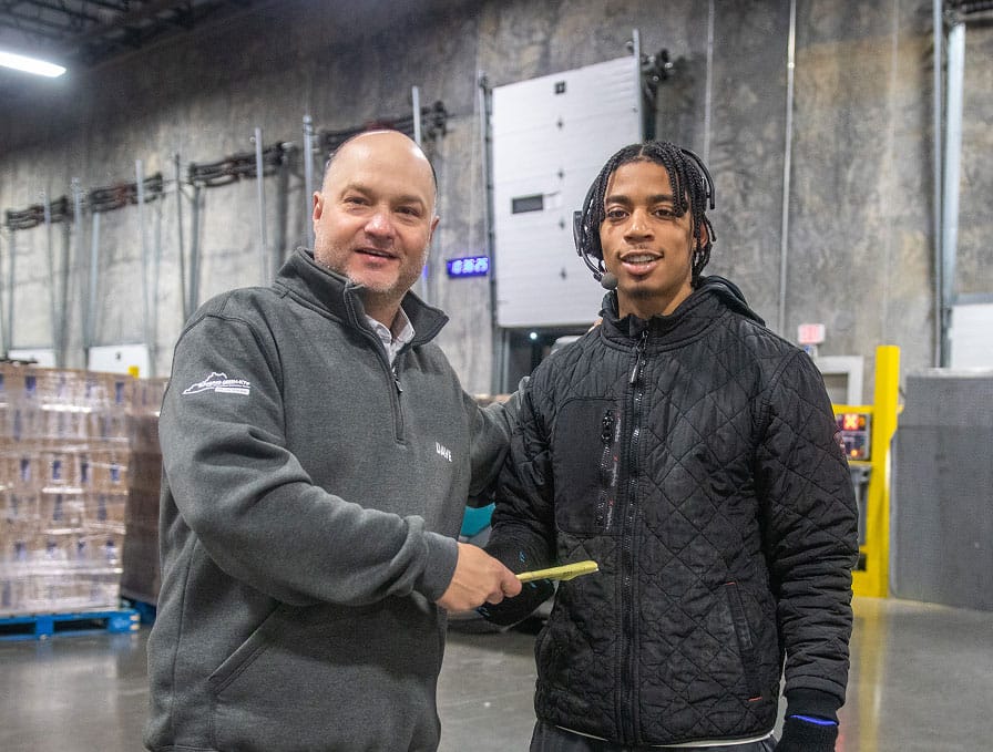 Two men stand in a warehouse, smiling at the camera. One man hands a piece of paper or envelope to the other. Pallets are visible in the background.