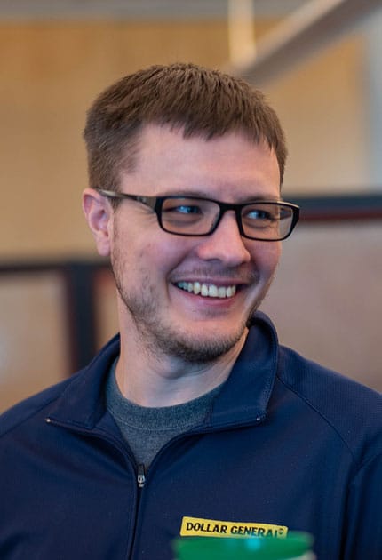 A man with short brown hair and glasses smiles while wearing a navy blue jacket with a “Dollar General” badge. The background is softly blurred.