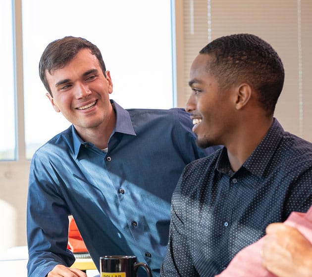 Two men in collared shirts are smiling and talking in an office setting, with a window and blinds in the background. One is holding a coffee mug.