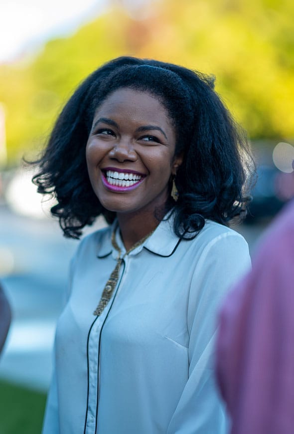 A woman with shoulder-length black hair and a bright smile wears a white blouse with black trim, standing outdoors with a blurred green background.