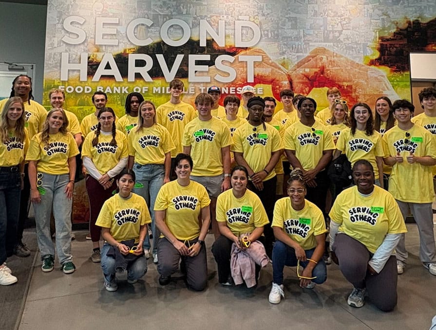 A group of people wearing matching yellow “Serving Others” shirts pose together in front of a “Second Harvest Food Bank of Middle Tennessee” sign.