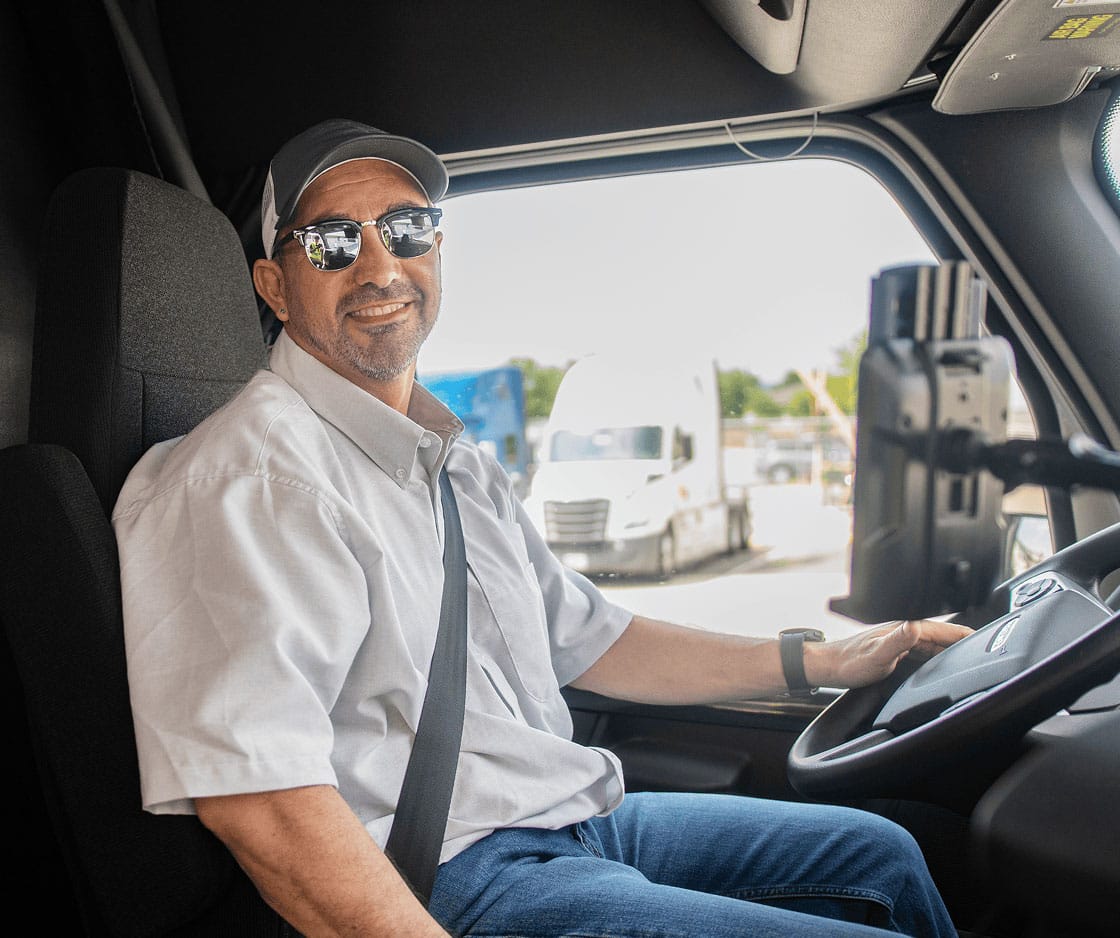 A smiling man wearing sunglasses and a cap sits in the driver’s seat of a truck, holding the steering wheel, with trucks visible outside the window.