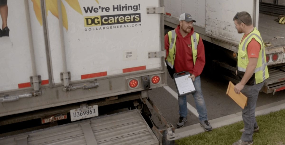 Two workers in safety vests stand behind a Dollar General truck. One holds a clipboard, and the other holds an envelope. A “We’re Hiring! DG careers” sign is on the truck.
