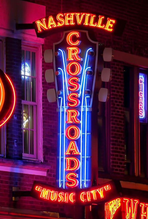 A neon sign shaped like a guitar reads “Nashville Crossroads Music City” in red, blue, and yellow lights, mounted on a brick building at night.