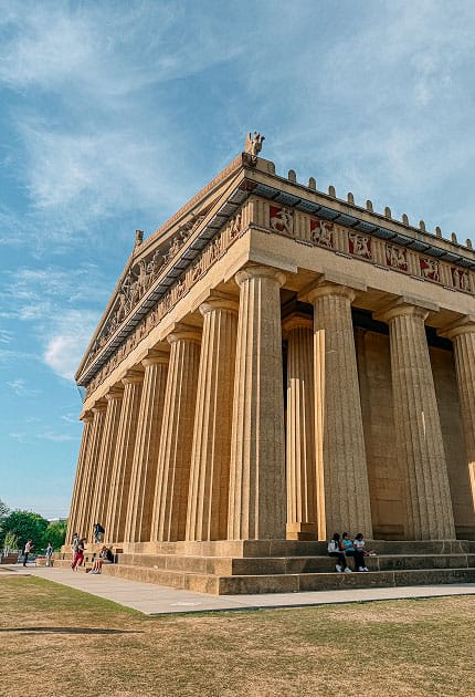 A large Parthenon replica with tall columns and detailed frieze under a blue sky. People are sitting on the steps and walking nearby. Grass and sidewalk are visible in the foreground.