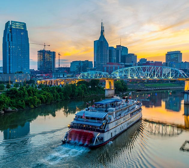 A riverboat cruises on a calm river at sunset, with a city skyline, tall buildings, and a lit bridge in the background.