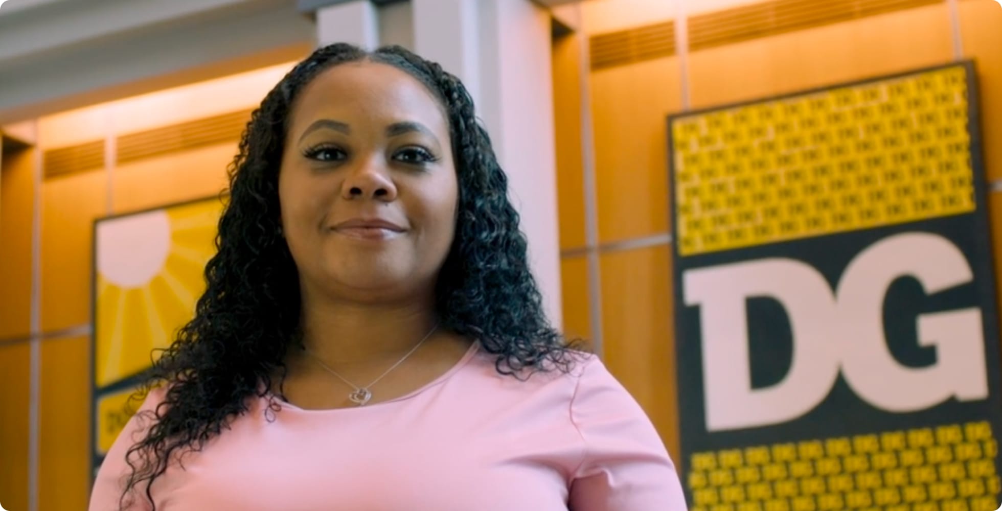 A woman with curly hair wearing a light pink top stands indoors, smiling slightly. Behind her are yellow and black signs, including one with large white letters “DG” on it.