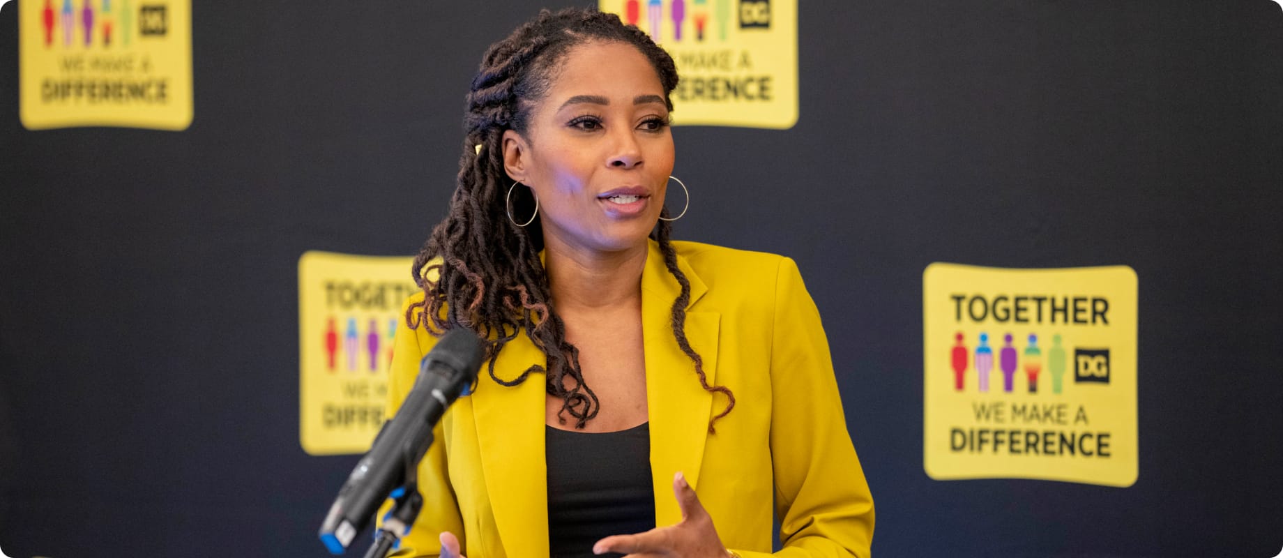 A woman in a yellow blazer speaks at a podium. Behind her are signs that read “TOGETHER WE MAKE A DIFFERENCE” with stylized people icons on a dark background.