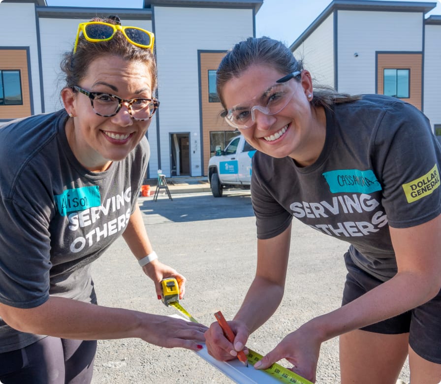 Two women wearing safety glasses and “Serving Others” shirts measure and mark a board outside, smiling at the camera. They appear to be volunteering at a building site with houses in the background.