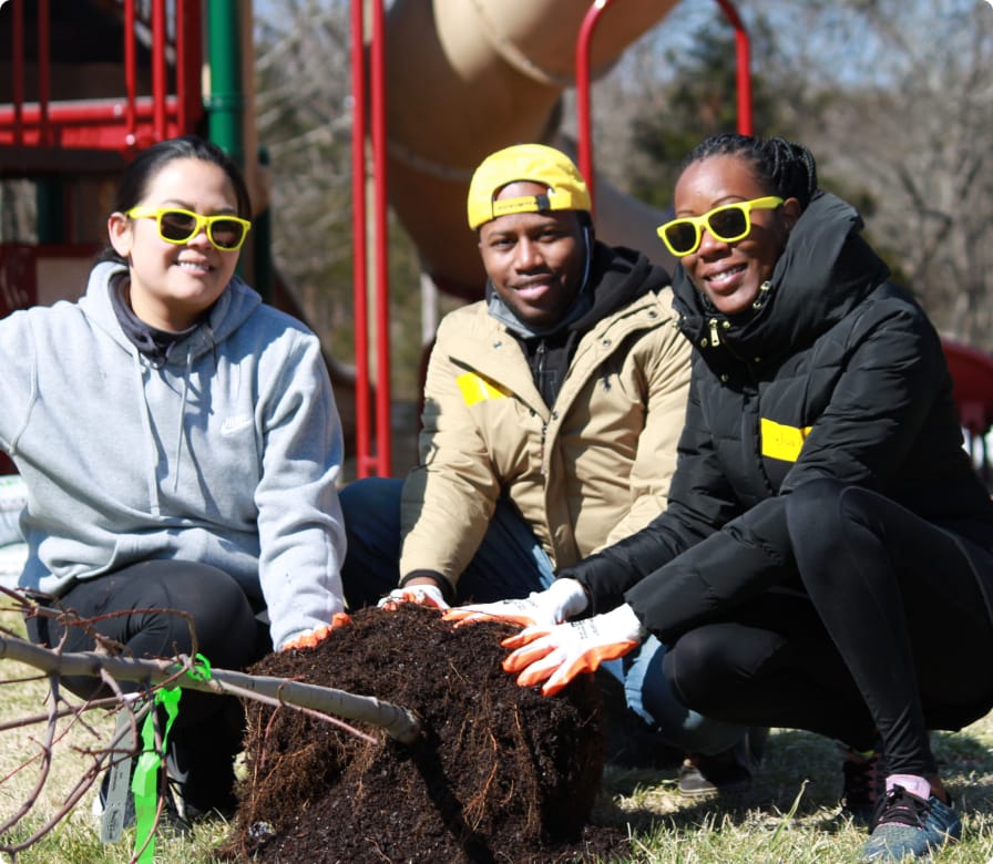 Three people wearing yellow sunglasses kneel outdoors, smiling and posing next to a tree sapling with exposed roots. They wear jackets and gloves, suggesting cool weather, with a playground visible in the background.