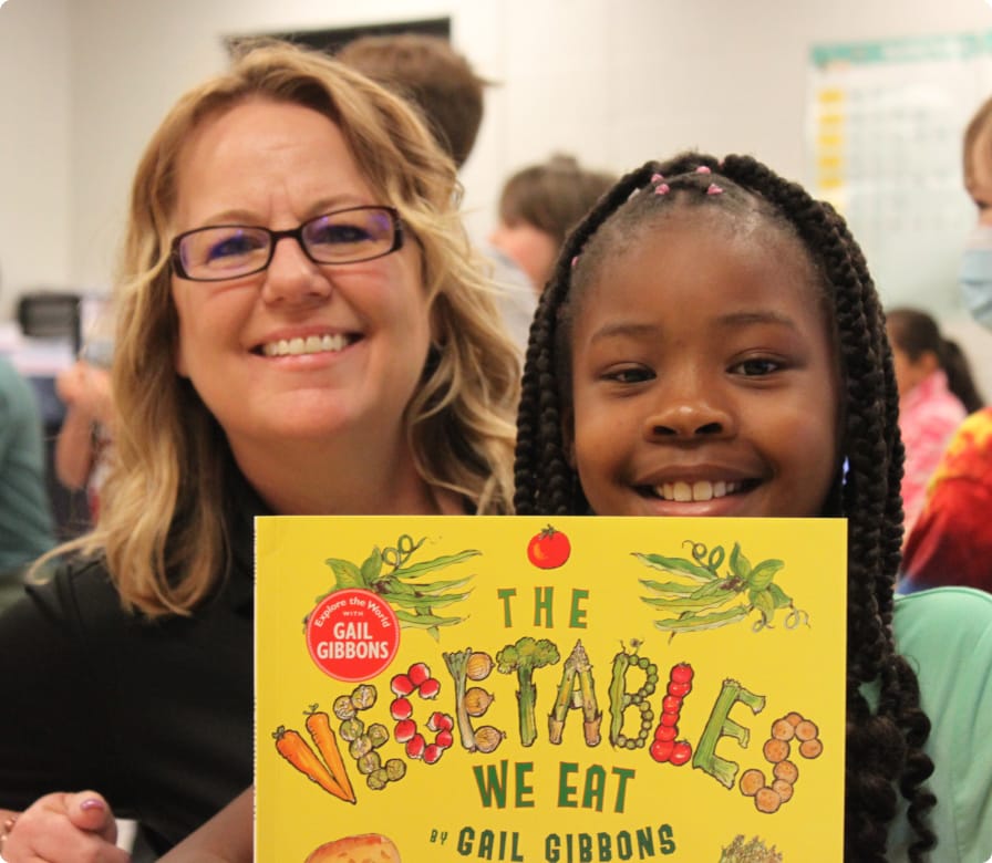 A smiling woman and girl hold up a colorful book titled “The Vegetables We Eat” by Gail Gibbons in a classroom setting.