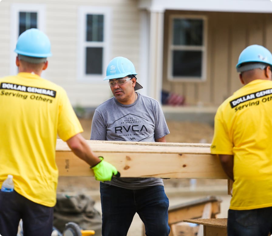 Three people wearing hard hats and gloves work together to carry a large wooden beam at a construction site. Two wear yellow shirts with “Dollar General Serving Others” on the back. A house is visible in the background.