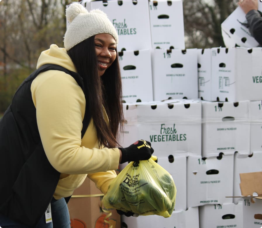 A smiling woman in a yellow hoodie and white beanie holds a bag of vegetables in front of stacked boxes labeled “Fresh Vegetables” outdoors.