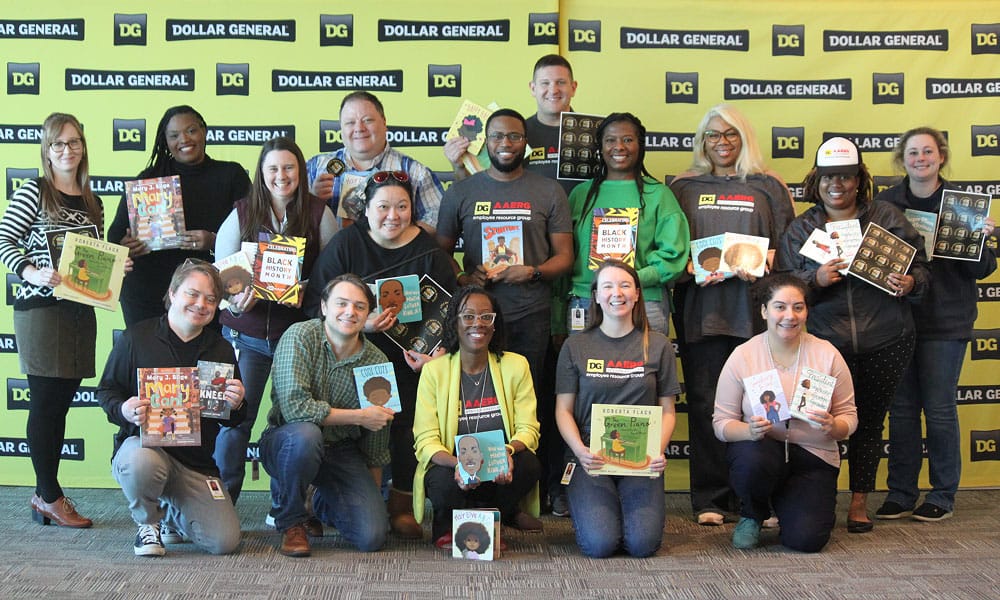 A diverse group of people pose and smile while holding books in front of a yellow Dollar General backdrop. Some wear Dollar General shirts and hats, and everyone is facing the camera.