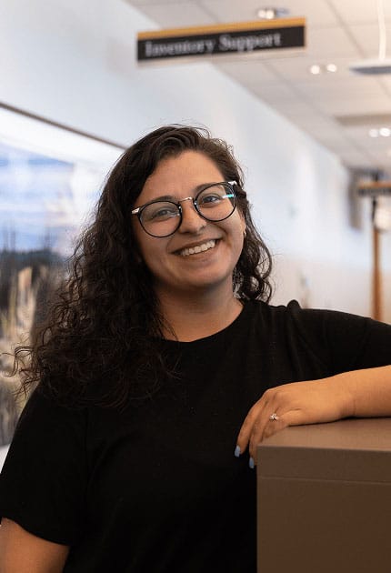 A woman with curly hair and glasses smiles while leaning on a counter. She is wearing a black shirt and standing indoors, with a sign reading “Inventory Support” visible in the background.