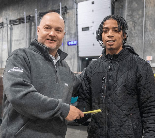 Two men standing indoors, smiling at the camera. One man, wearing a gray zip-up, is holding a yellow paper while placing his arm around the other man, who wears a black quilted jacket and has braided hair.