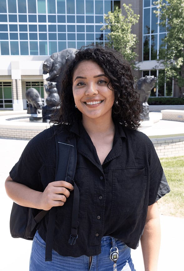 A young woman with curly hair smiles while standing outside a building, wearing a black shirt and backpack. Bear statues and green trees are visible in the background.