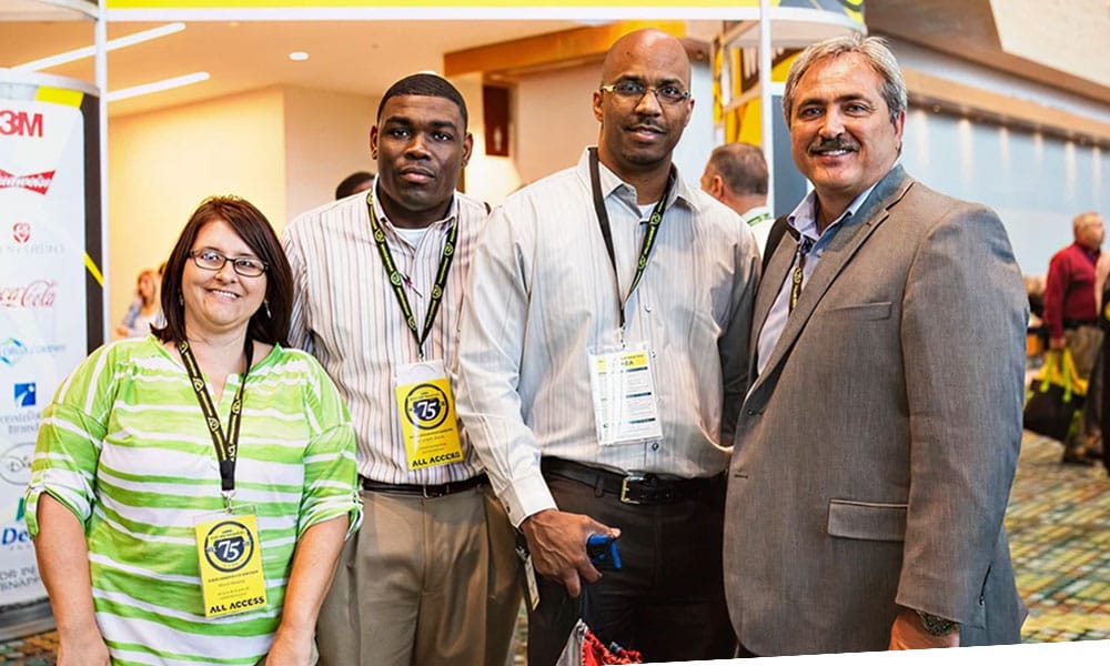 Four people, three men and one woman, stand together at a conference or event, wearing badges and business casual attire, smiling at the camera in a brightly lit indoor setting.