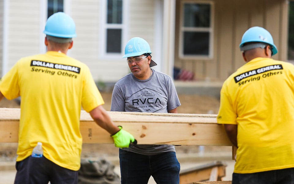 Three men wearing hard hats lift wooden beams at a construction site. Two wear yellow “Dollar General Serving Others” shirts, while the third wears a gray RVCA shirt. A house under construction is in the background.
