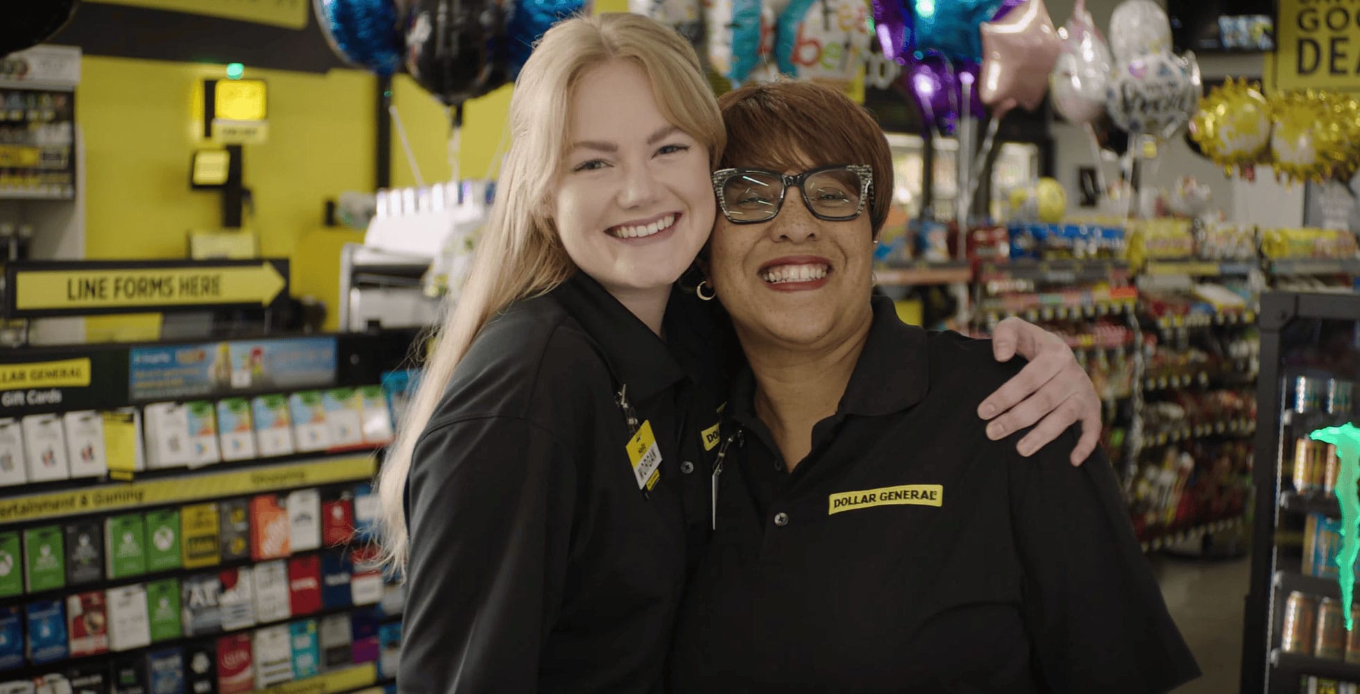 Two Dollar General employees wearing black uniforms smile and hug each other inside the brightly lit store, surrounded by colorful balloons and various products on display shelves.