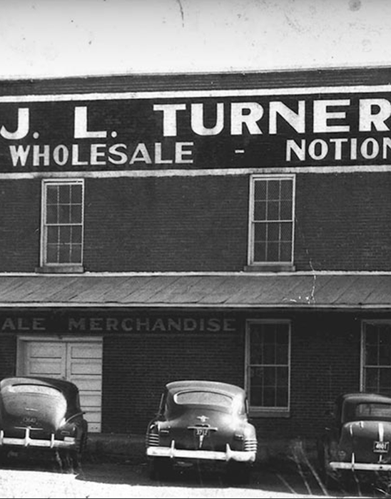 Black-and-white photo of a brick building labeled “J.L. TURNER WHOLESALE - NOTIONS.” Four classic cars are parked in front. The lower sign reads “MERCHANDISE.” The scene appears to be from the mid-20th century.
