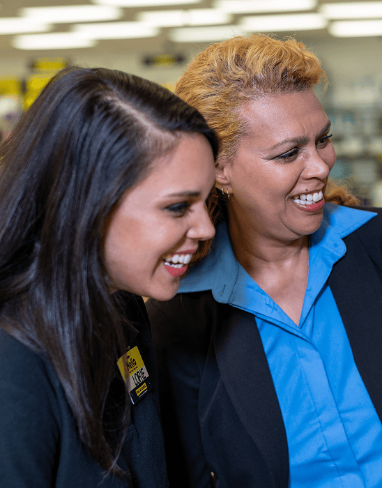 Two women smiling and standing close together indoors. One wears a yellow name tag and a black blazer, while the other wears a blue shirt and black blazer. Shelves with products are blurred in the background.