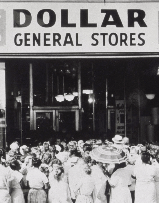 A large crowd of people stands outside a Dollar General Store, looking toward the entrance under a large sign. One person holds a patterned umbrella. The image is black and white.