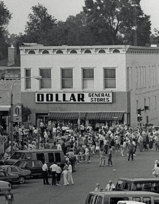 A large crowd gathers in front of a Dollar General Store on a city street; many people stand on the sidewalk and street, with numerous vehicles parked nearby.