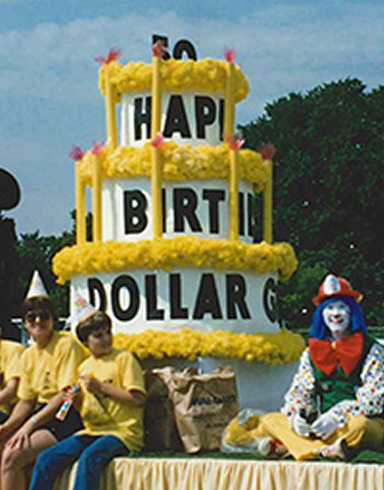 People in yellow shirts sit by a large, three-tiered cake float with “HAPPY BIRTHDAY DOLLAR GENERAL” on it. A clown in bright clothes and a pointed hat sits on the right side of the float.