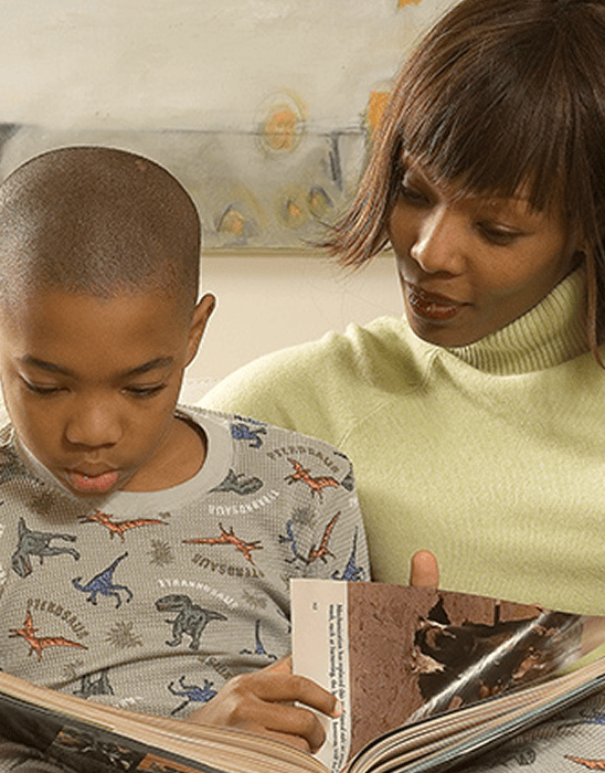 A woman and a young boy sit closely together reading a book. The boy wears dinosaur pajamas and focuses on the pages, while the woman looks at him, smiling gently. A painting is visible in the background.