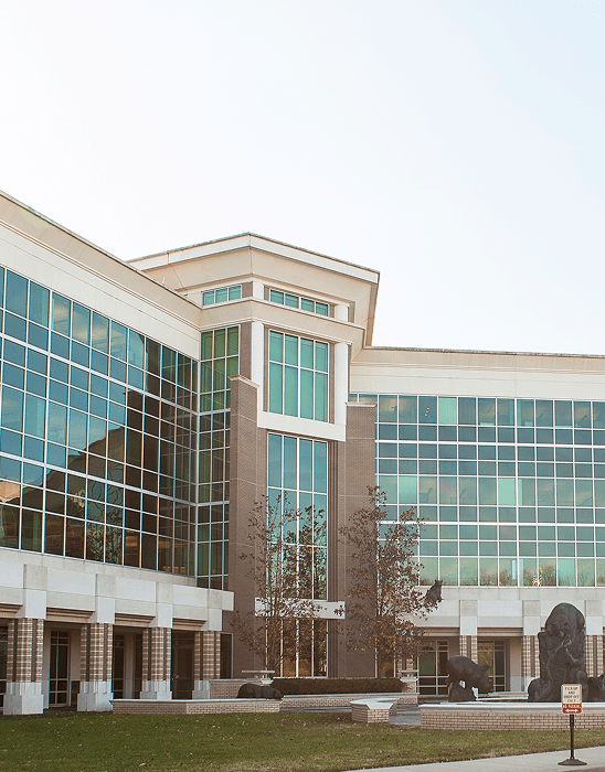 Modern building with large glass windows and brick accents, featuring a courtyard with trees, sculptures, and a green lawn in the foreground. The sky is clear and bright.