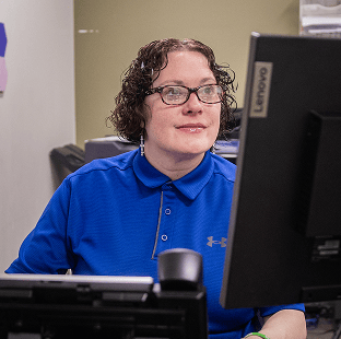 A person with curly hair and glasses, wearing a blue polo shirt, sits at a desk working on a Lenovo computer. The background shows office equipment and shelves.
