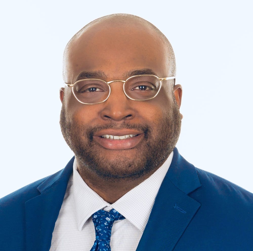 Man wearing glasses, a blue suit, white shirt, and blue patterned tie, smiling at the camera against a light background.