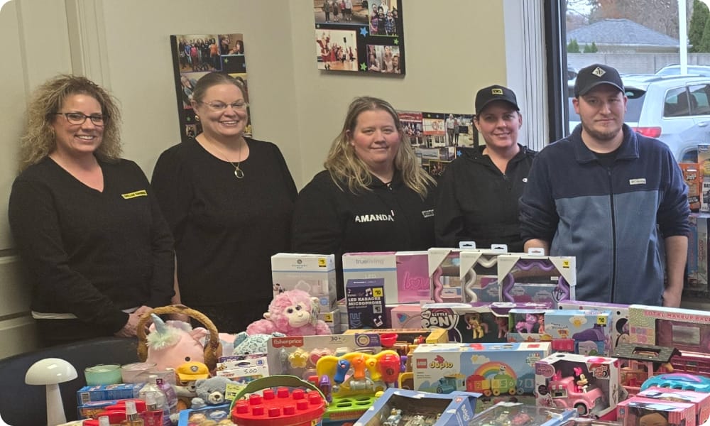 Five people stand behind a table filled with boxed toys, plush animals, and games. They are smiling in an office setting, with photos displayed on the wall behind them and windows showing the outdoors.
