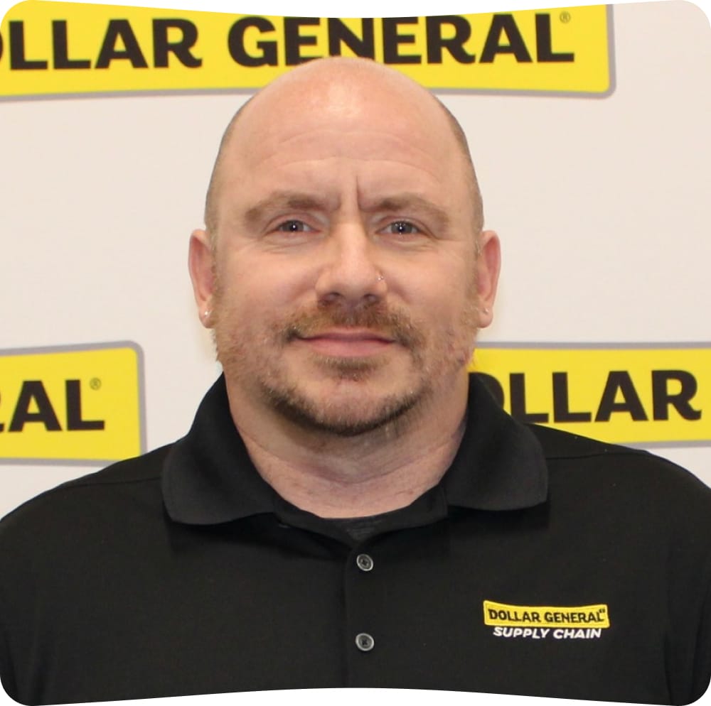 A person in a black “Dollar General Supply Chain” polo shirt stands in front of a Dollar General sign, smiling slightly at the camera.