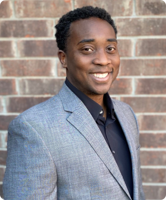 A man in a gray suit jacket and black shirt smiles while standing in front of a brick wall.