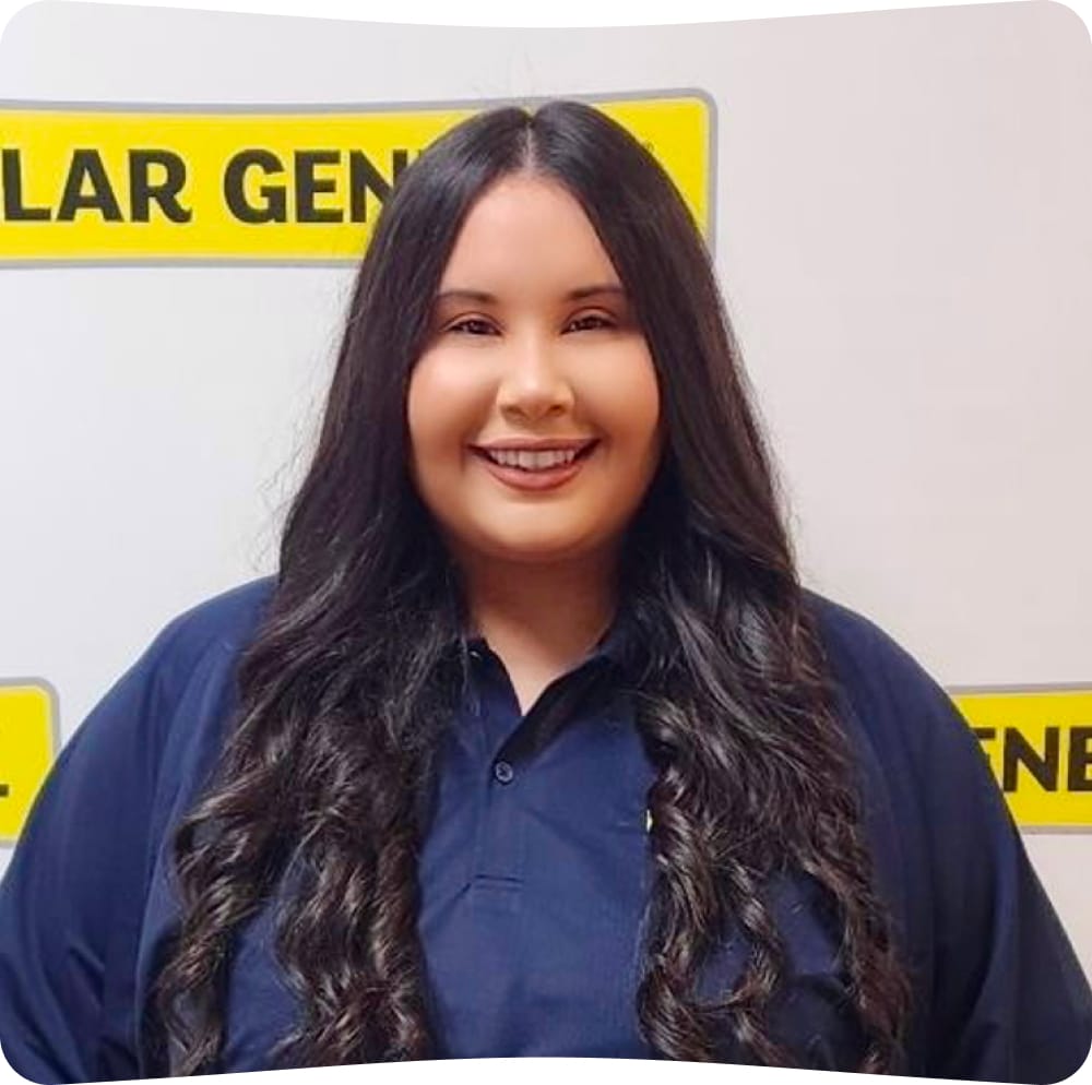 Woman with long dark hair, wearing a navy blue collared shirt, smiles in front of a white wall with a partially visible yellow and black “General” sign.