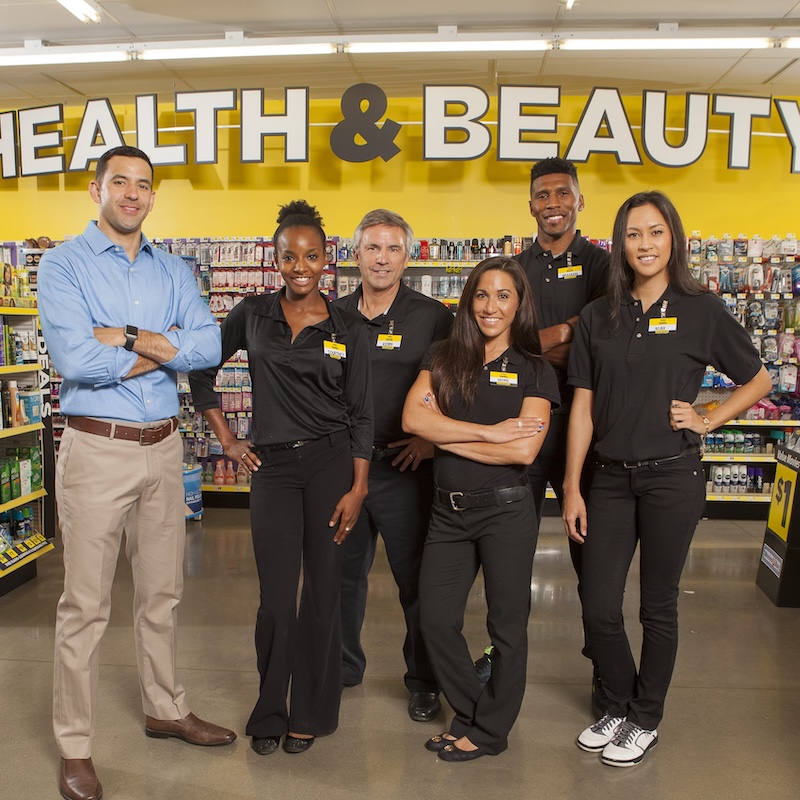 Five store employees stand smiling in front of a “Health & Beauty” section at a retail store, wearing black or blue uniforms with name tags, surrounded by shelves of products.